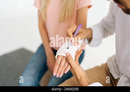 Gros plan vue de dessus d'un ami mâle méconnaissable écrivant l'inscription sur le casting de la femme avec une main cassée enveloppée dans un bandage en plâtre. Banque D'Images