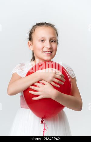 Portrait sourire fille embrassant rouge ballon avec les deux mains, regardant l'appareil photo. Jolie fille joyeuse 10 ans jouant avec ballon. Prise de vue en studio Banque D'Images