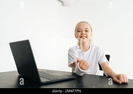 Bonne petite fille blonde montrant le pouce vers le haut mouvement avec main cassée enveloppé dans le bandage en plâtre blanc assis à la table avec ordinateur portable souriant regarder Banque D'Images