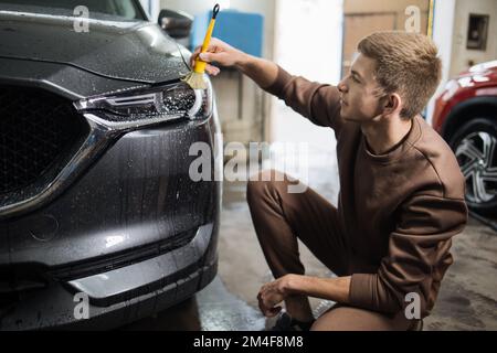 Lavage de voiture, concept de détail. Vue latérale de l'homme caucasien travailleur de lavage de voiture nettoyage des phares de voiture grise de luxe à l'aide d'une bouteille de pulvérisation verte. Banque D'Images