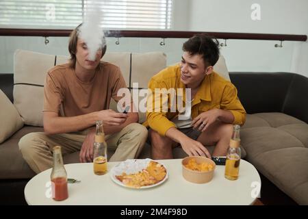 Les jeunes hommes fument des cigarettes électroniques à la maison lorsqu'ils ont une pizza avec de la bière Banque D'Images