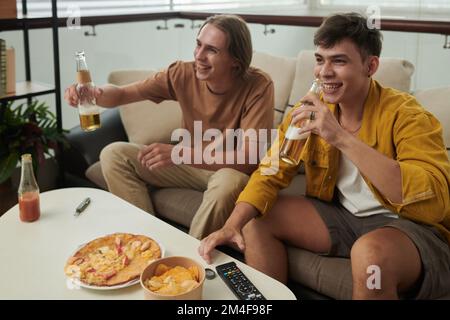 Des jeunes hommes heureux qui boivent de la bière et mangent de la pizza lorsqu'ils regardent un match à la télévision à la maison Banque D'Images