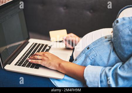 Shopping pour le berceau parfait de bébé confortablement de la maison. une femme enceinte utilisant un ordinateur portable et une carte de crédit sur le canapé à la maison. Banque D'Images