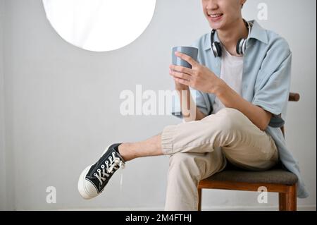 Le jeune homme asiatique heureux s'assoit sur une chaise dans un salon très lumineux, aime siroter un café le matin. image rognée Banque D'Images