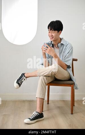 Un jeune homme asiatique heureux et séduisant assis sur une chaise dans un salon très lumineux, aime siroter un café le matin. Banque D'Images