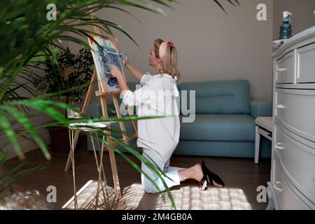 Une artiste féminine tire avec enthousiasme sur la toile avec ses mains les doigts et la brosse sur le sol de la maison Banque D'Images