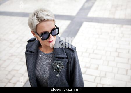 Portrait d'une femme mûre à la mode avec des cheveux blonds courts portant un élégant manteau en laine grise avec des revers et des bretelles, une broche d'accessoires et le soleil Banque D'Images