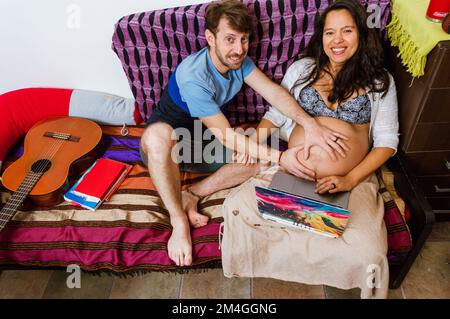 Homme adulte caucasien touchant le ventre de sa femme enceinte assis ensemble sur le canapé et regardant l'appareil photo, tout en utilisant l'ordinateur portable. Banque D'Images