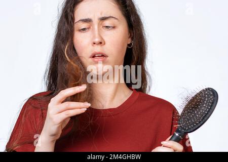 Gros plan portrait d'une femme brunette attristée tient un peigne avec les cheveux tombant et regarde les bouts de ses cheveux. Arrière-plan blanc. Concept de cheveux Banque D'Images