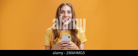 Prise de vue à la taille d'une jeune femme mignonne, amusante et excitée, avec une coiffure ondulée, souriante et étonnamment étonne en réaction à une tenue de smartphone impressionnante Banque D'Images