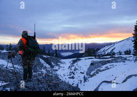 Chasseur avec carabine vêtue de camo se dresse sur le bord d'une crête de montagne enneigée à la recherche de cerfs au lever du soleil dans le Wyoming Banque D'Images