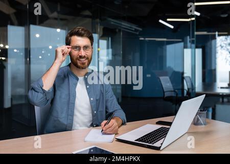 Portrait d'un homme d'affaires prospère derrière un travail papier, d'un homme en chemise souriant et regardant l'appareil photo, d'un responsable financier au bureau utilisant un ordinateur portable au travail. Banque D'Images