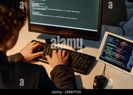 Vue arrière d'un jeune programmeur utilisant un clavier pour taper des codes pour un nouveau logiciel pendant son travail au bureau jusqu'à tard dans la nuit Banque D'Images