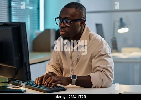 Un sérieux programmeur américain africain de lunettes dactylographiant sur ordinateur pour développer un nouveau programme, il travaille des heures supplémentaires au bureau Banque D'Images
