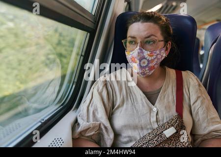 Une fille en lunettes et un masque médical qui monte dans un train et regarde par la fenêtre Banque D'Images