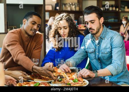 Divers employés d'équipe ayant une pizza ensemble dans un café. Un homme africain prenant une tranche de pizza de l'assiette. Le personnel partage les repas. Profiter de la pause déjeuner. Banque D'Images