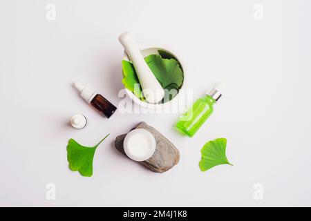 Feuilles de ginkgo vert frais, mortier et pilon, bouteille de sérum cosmétique et pot à crème sur fond blanc. Concept de cosmétiques naturels faits à la main. Vue de dessus Banque D'Images