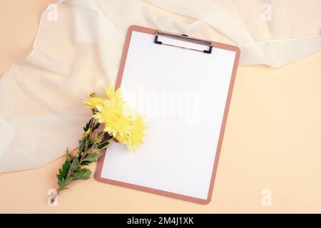 Presse-papiers avec papier vierge et chrysanthèmes jaunes sur fond beige. Vue de dessus, plan d'agencement, maquette Banque D'Images
