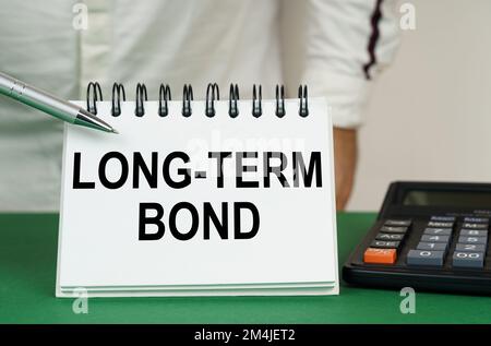 Concept d'entreprise. Sur une surface verte se trouve une calculatrice, un homme pointe avec un stylo vers un bloc-notes avec l'inscription - long-Term Bond Banque D'Images