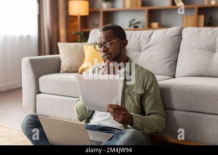 Pensive sérieux adulte noir homme dans les lunettes utilise l'ordinateur, travaille avec des documents, assis sur le sol dans le salon Banque D'Images