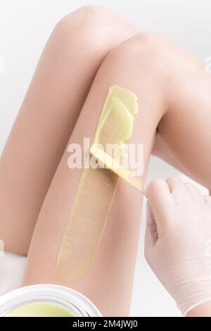 Esthéticienne en gants appliquant de la cire chaude verte sur la jambe de femme mince à l'aide d'une spatule. Épilation à la cire chaude dans un salon de beauté Banque D'Images