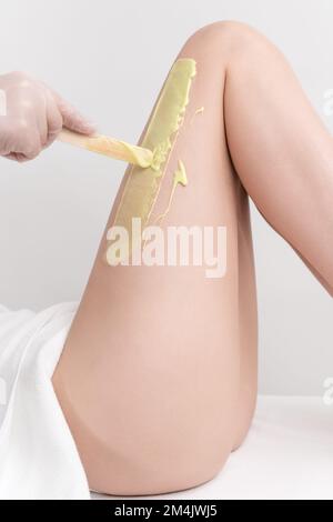 Main dans le gant application de cire chaude verte sur la jambe femme mince à l'aide d'une spatule. Procédure d'épilation à la cire chaude dans un salon de beauté Banque D'Images