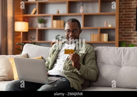 Sourire adulte noir gars dans les lunettes regarde la carte de crédit, utilise l'ordinateur pour les achats en ligne, vérifie le compte bancaire Banque D'Images