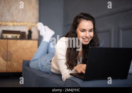 Bonne femme caucasienne utilisant un ordinateur portable, travaillant ou étudiant en ligne, allongé sur un canapé à la maison, espace libre Banque D'Images