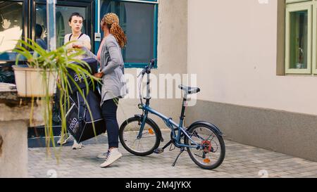 Un service de livraison express de nourriture livrant le déjeuner dans un sac en papier, donnant la commande de fastfood à la porte d'entrée. Client souriant prenant un repas à emporter au restaurant à l'extérieur, femme avec forfait à vélo. Banque D'Images