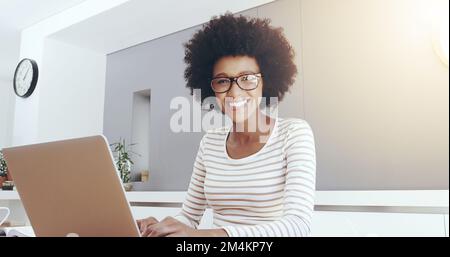 Elle est très confiante dans son travail. Portrait d'une jeune femme gaie qui tape et travaille sur un ordinateur portable tout en étant assise à côté d'une table à la maison. Banque D'Images
