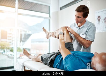 Je peux ressentir de la douleur autour de mon genou. un jeune kinésithérapeute masculin aidant un client avec des exercices de jambe qui se trouvent sur un lit. Banque D'Images