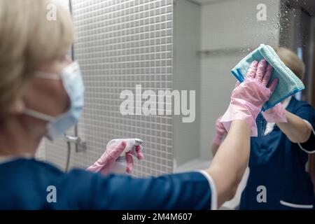 La femme de chambre dans le masque nettoie le miroir dans la salle de bains de l'hôtel en vaporisant du détergent sur la surface Banque D'Images