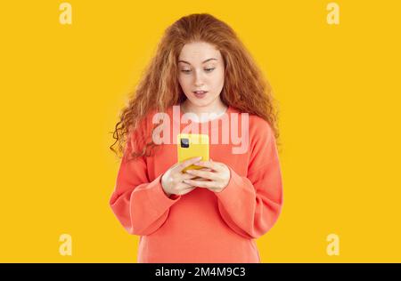 Frustré et stupéfait fille de tête rouge avec des cheveux bouclés regardant l'écran de smartphone sur fond jaune. Banque D'Images