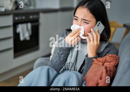 Portrait d'une jeune femme coréenne malade se sentant malade, éternuant et tenant une serviette, restant malade à la maison, attrapée par le froid. Parler sur un téléphone portable Banque D'Images