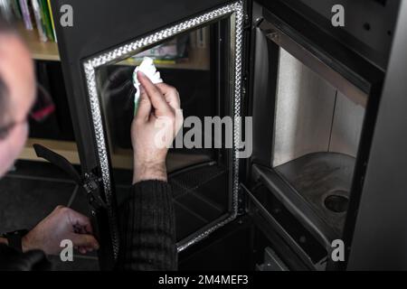 Homme nettoyant la cendre sur le verre d'une pastille ou d'un poêle à bois avec une serviette en papier et de l'eau chaude Banque D'Images