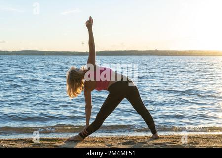 Vue arrière d'une femme inclinée latéralement. Une femme pratiquant le yoga au bord du lac en été. Méditation. Yoga sur la côte. Sports d'été Banque D'Images