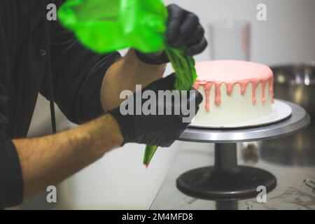 le chef cuisinier prépare des gâteaux dégoulinés sur un gâteau blanc givré pour la fête d'anniversaire de bébé fille au laboratoire de cuisine avec garniture rose sur le sac à passepoil Banque D'Images
