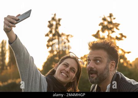 Mignon jeune femme caucasienne tenir smartphone prendre selfie avec beau homme dehors dans la nature avec le visage stupide. Père et fille famille amusant temps ensemble Banque D'Images