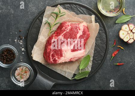 Steak de bœuf cru. Steak de rouleau Chuck frais cru avec romarin, sel et poivre sur planche à découper sur fond de béton foncé. Steak de bœuf cru et épices pour Banque D'Images