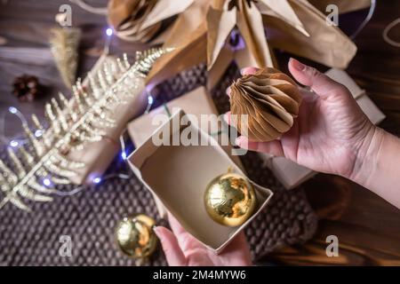 Main tenant un jouet fait main en papier artisanal. Concept zéro déchet. Emballage des cadeaux de Noël. Boîte-cadeau en papier avec jouets dorés pour arbres de Noël. Banque D'Images