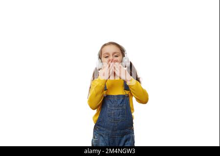 Gai charmante petite fille soufflant des baisers d'air, écoutant de la musique sur des écouteurs, isolé sur fond blanc Banque D'Images