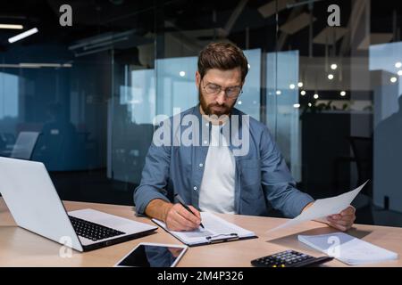 Un sérieux jeune homme comptable, financier, analyste, auditeur est assis dans le bureau à la table. Fonctionne avec les documents. écrit et calcule le budget, les finances. Banque D'Images