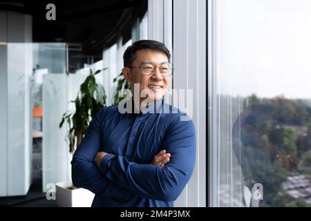 Portrait d'un homme d'affaires souriant réussi, d'un homme asiatique avec des mains froissées dans une chemise décontractée donnant sur la fenêtre, d'un homme mature dans des lunettes travaillant à l'intérieur d'un bureau moderne. Banque D'Images