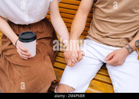 Prise de vue courte en grand angle d'un jeune couple heureux et méconnaissable amoureux, en tenant les mains sur le banc et en buvant un café à emporter tout en appréciant le temps Banque D'Images