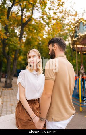 Photo verticale d'un jeune couple heureux et calme, amoureux de la main dans le parc d'attractions, sur fond de carrousel, le jour de l'été. Banque D'Images