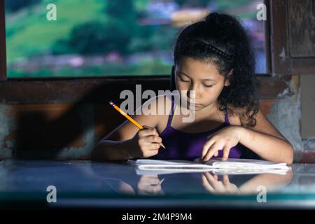 latina fille, brunette étudiant. petite fille avec un crayon jaune faisant ses devoirs, s'appuyant sur une table en verre. Banque D'Images