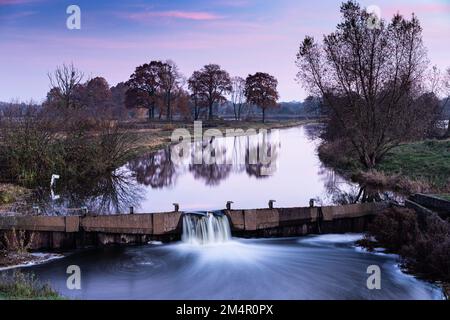 Le soleil levant baigne la rivière, les arbres, le ciel, les nuages et le déversoir sous une lumière rouge. Banque D'Images