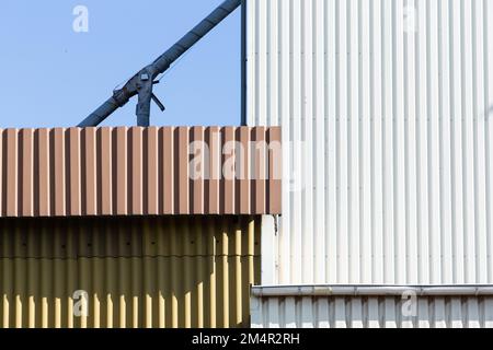 Les façades métalliques d'un bâtiment industriel blanc, marron et vert sont décalées et apparaissent abstraites par rapport à un ciel bleu. Banque D'Images