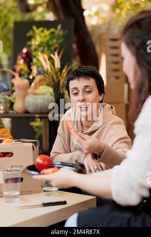 Les femmes goûtent une tranche de tomate sur le stand du marché agricole local, discutant avec le propriétaire d'une petite entreprise des produits frais. Client souriant et appréciant le goût de fruits et légumes biologiques. Banque D'Images