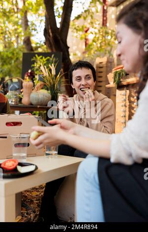 Une jeune femme heureuse qui apprécie la dégustation de fruits biologiques et de légumes sur le marché alimentaire local le jour ensoleillé de l'automne. Agricultrice assise à table avec le consommateur, offrant de déguster des produits maison Banque D'Images